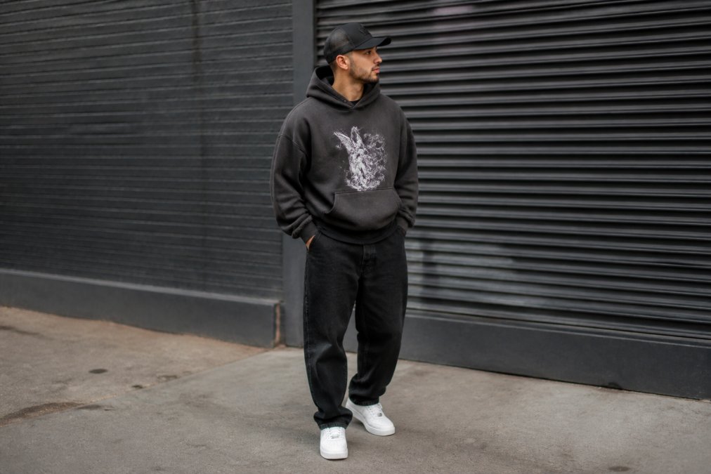Jongvolwassene in een oversized HTT-hoodie met T-shirt met print, witte sneakers en een gestructureerde pet in een stedelijke omgeving met natuurlijk daglicht, casual streetwear-styling
