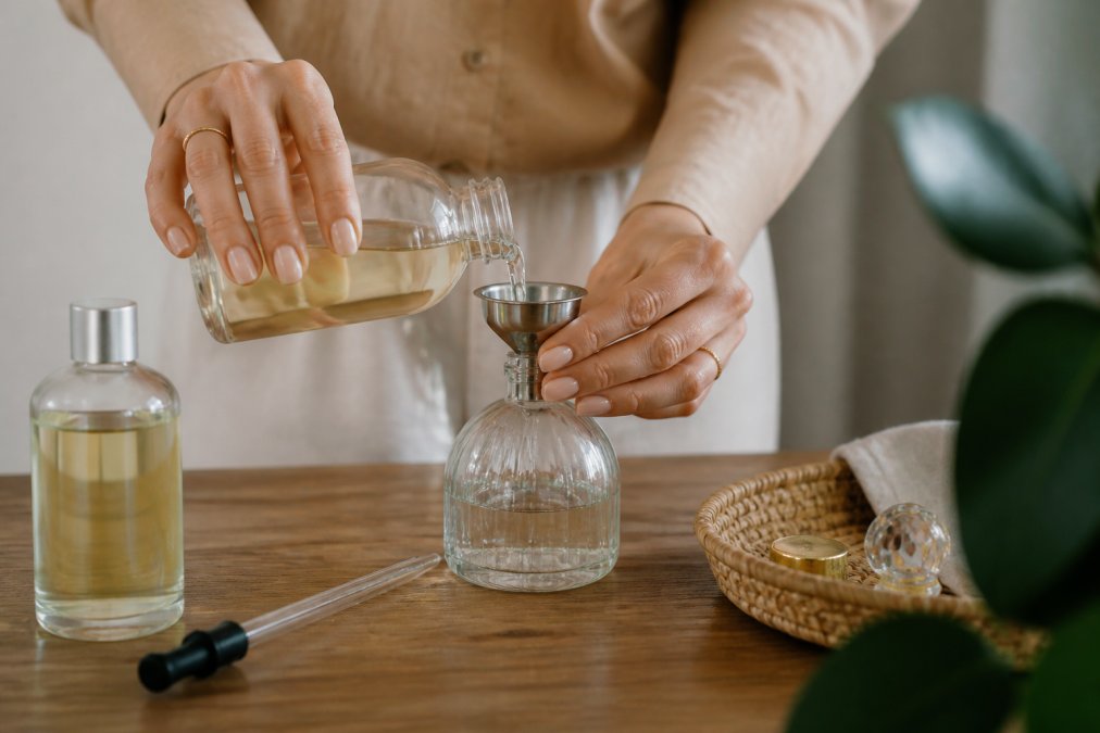 Close-up van handen die een glazen parfumfles met een trechter voorzichtig bijvullen op een houten tafel, met een fles huisparfum en een pipet ernaast, warm natuurlijk licht