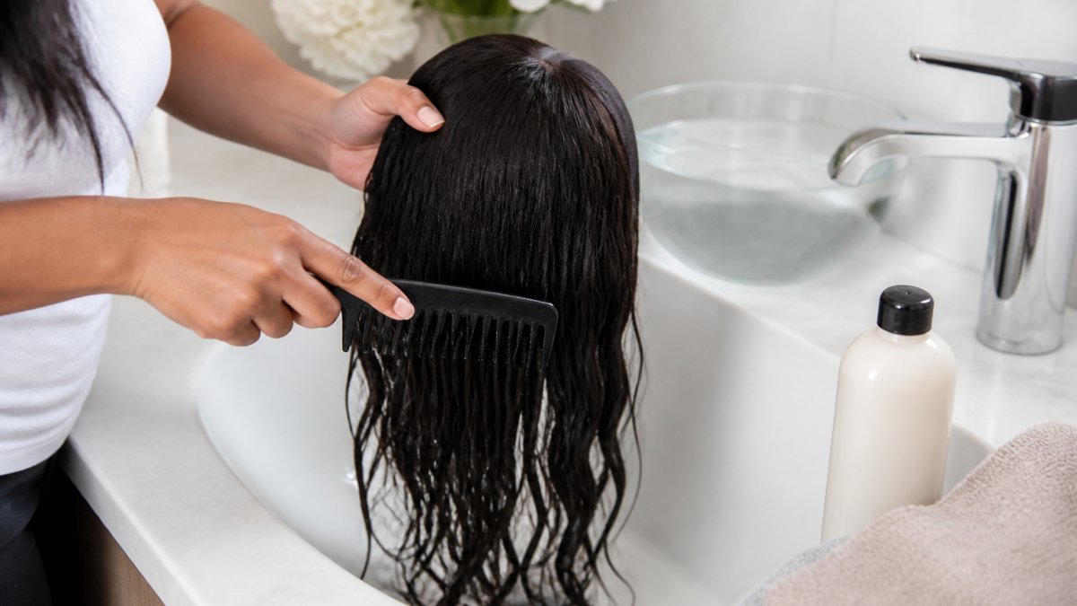 Close-up of a woman's hands gently combing a dark human hair wig with a wide-tooth comb over a bathroom sink, wig shampoo bottle and cool water visible, soft natural light