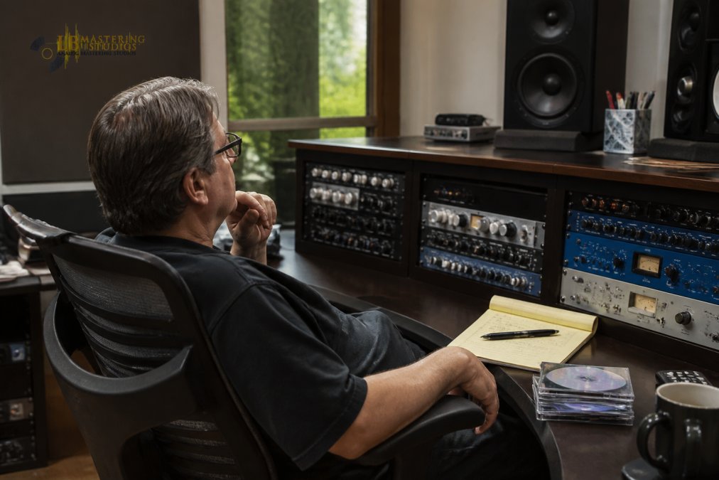 Recording artist or music producer reviewing a mastered track on professional studio monitors, leaning back in an ergonomic chair with handwritten notes and reference CDs visible on the desk, natural studio lighting illuminating their focused expression