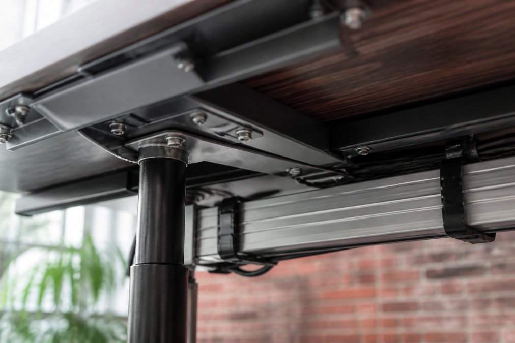 Close-up of metal desk frame joints, pneumatic lift cylinder, cable management clips, and aluminum support beam showing construction quality and durability