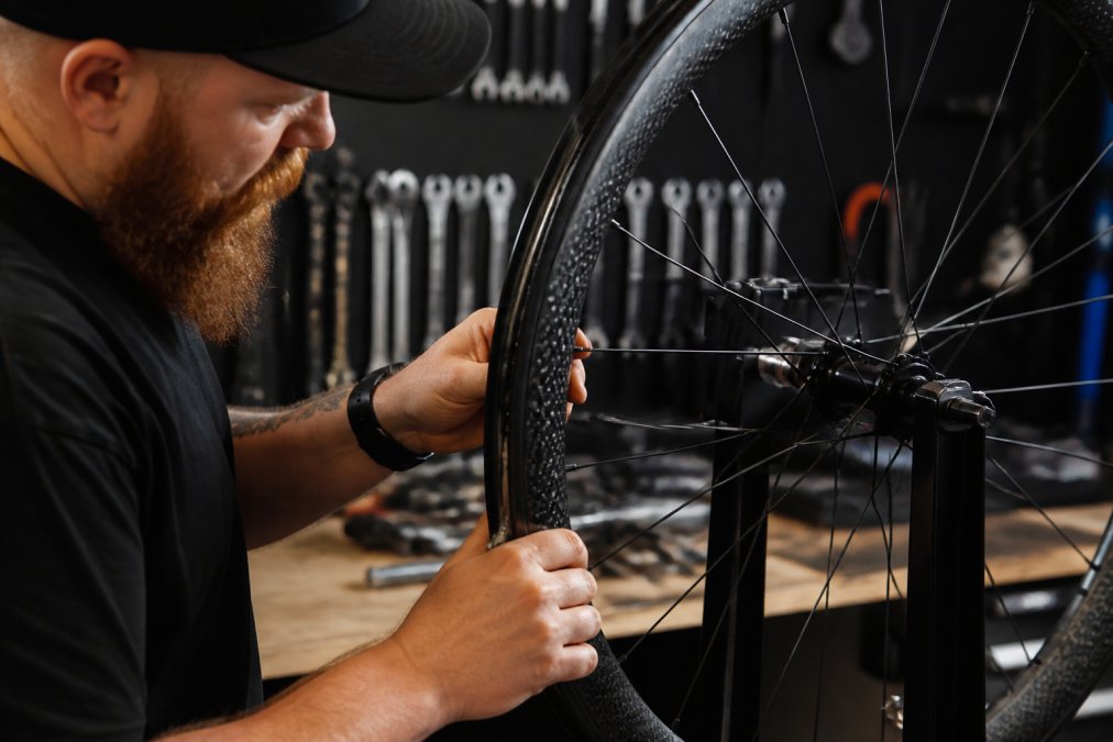 Close-up of a wheelbuilder's hands adjusting spoke tension on a carbon rim during final truing, with precision tools and workshop lighting visible in the background