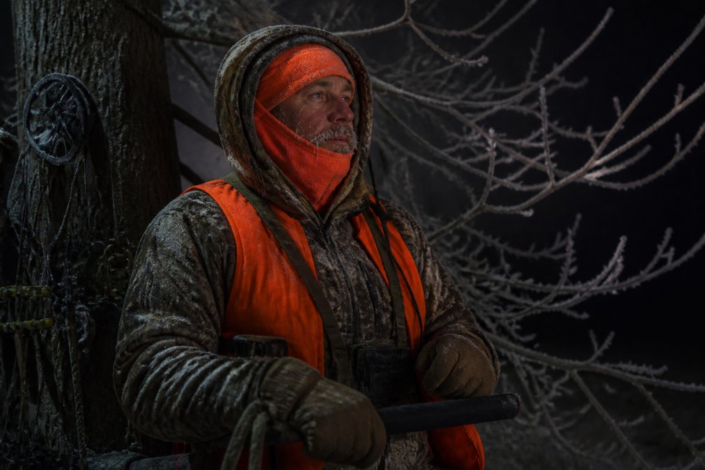 A hunter bundled in heavy insulated gear and blaze orange standing in a treestand during pre-dawn darkness with frost visible on branches and the sky still black, looking determined but exhausted