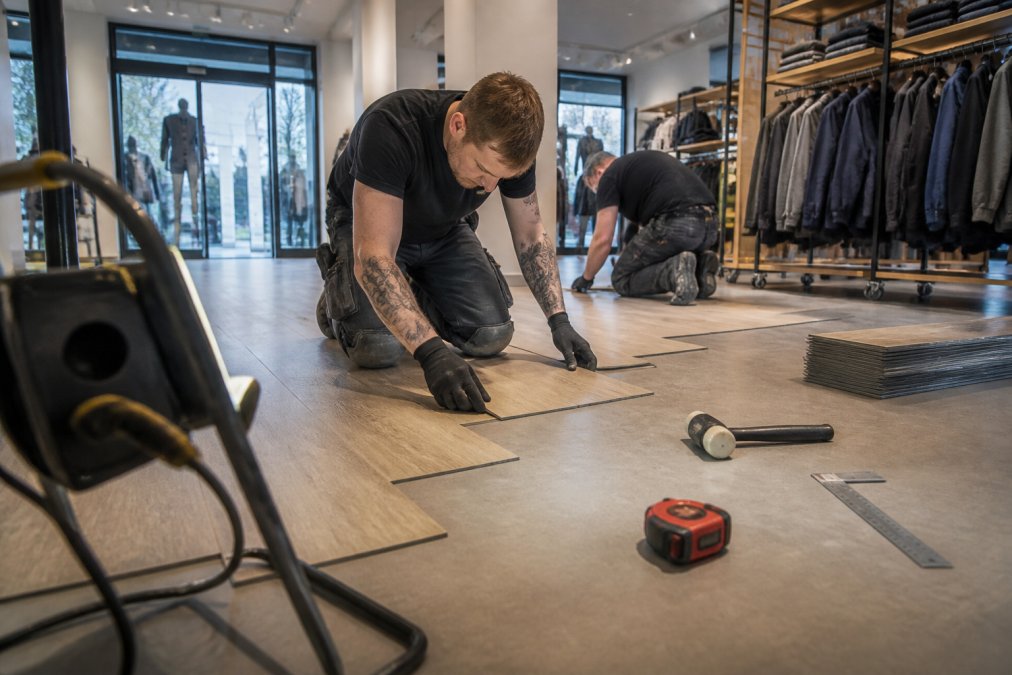 Håndværkere installerer klikgulve i en butik, tæt på gulvet med kliksystemet i aktion, værktøj og materialer synlige, arbejdslamper belyser området