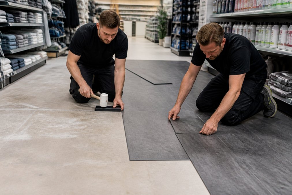 Hurtig lægning af gulv i butik: Guide til 2026 2 Two retail workers in a bright store aisle installing interlocking vinyl floor tiles over a level subfloor, one kneeling with a tapping block, the other aligning a tile edge, shelves with products in the background