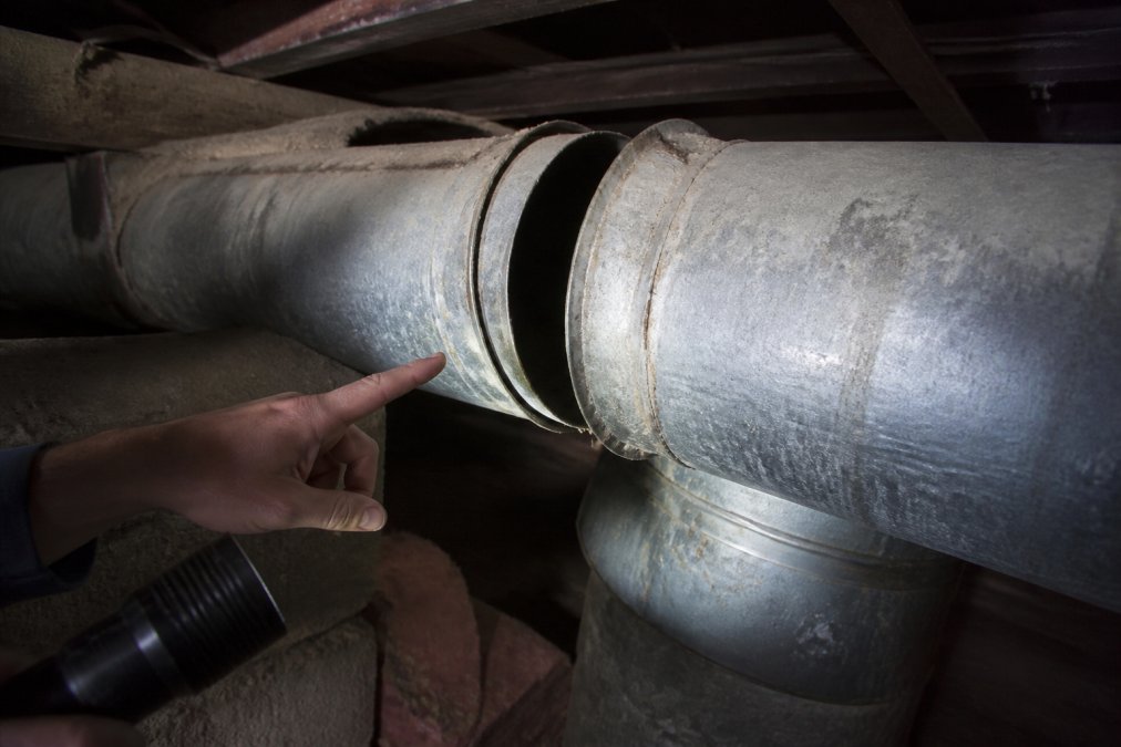 A technician's hand pointing to a corroded, disconnected duct joint in a dusty basement, a flashlight illuminating the gap and the worn metal edges