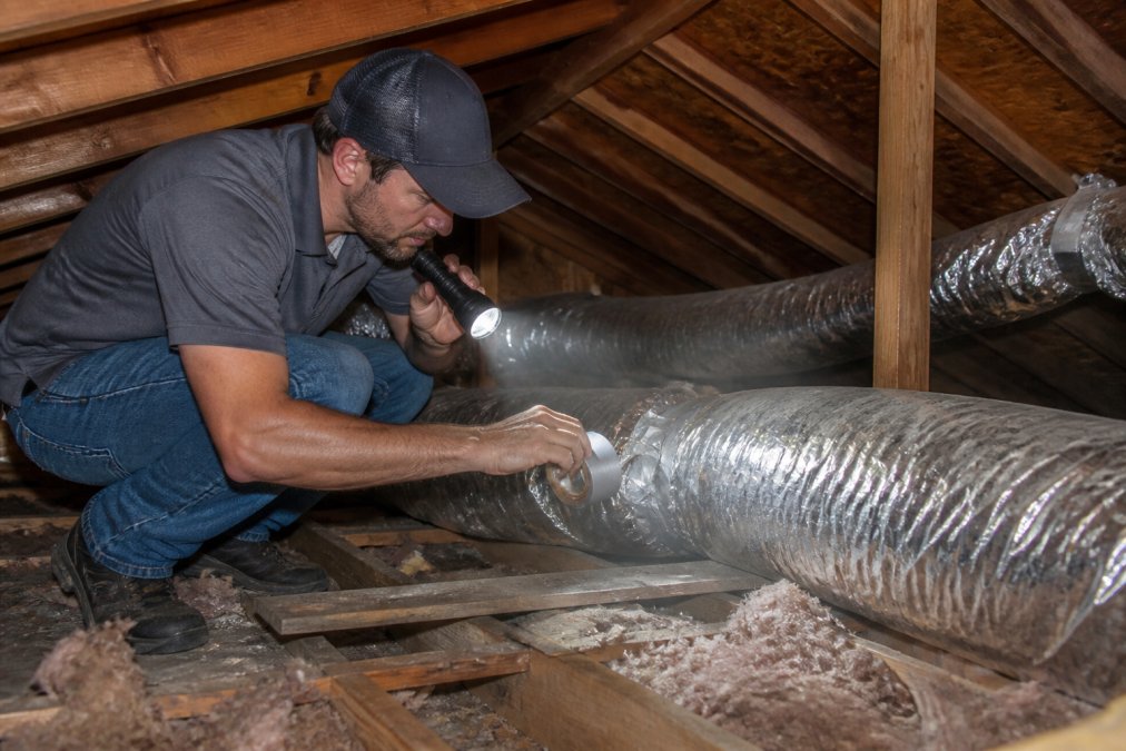 HVAC technician crouching in an attic space with a flashlight, inspecting flexible ductwork connections and sealing gaps with duct tape, dust visible in the beam of light