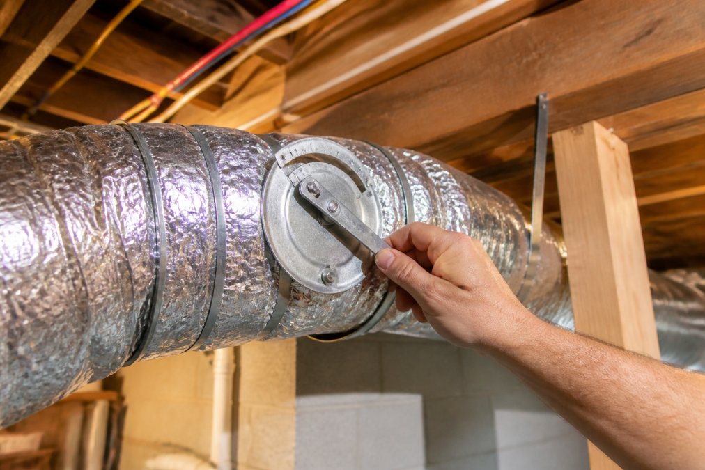Homeowner's hand adjusting a lever-style damper control on flexible ductwork in a basement, with the damper positioned at a 45-degree angle to partially restrict airflow