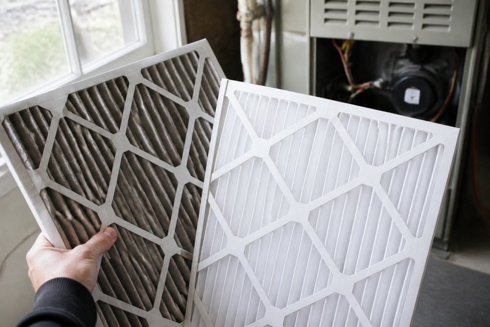 A homeowner's hand holding a dirty, gray HVAC air filter beside a fresh white replacement filter, with an open furnace compartment visible in the background and natural window light illuminating the contrast