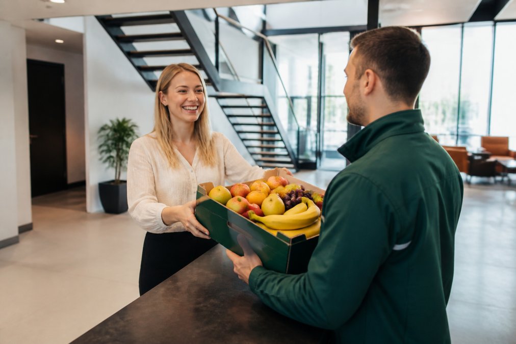 En smilende medarbejder modtager en kasse med frisk frugt fra en leverandør i uniform ved receptionen i et moderne kontor med dagslys
