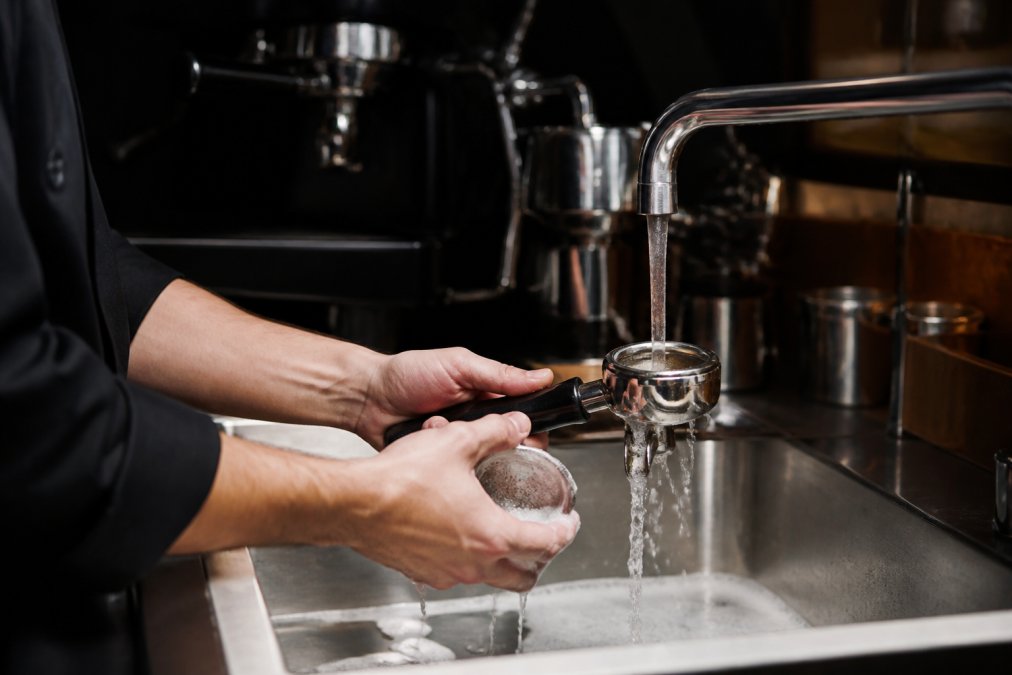 Barista, der rengør bryggehovedet og portafilter under rindende vand ved en køkkenvask med sæbeskum og vand