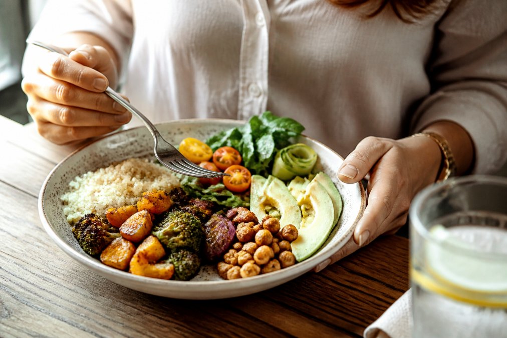 Nærbillede af en persons hænder, der holder en tallerken med farverige grøntsager, frugt og hele kornprodukter, mens personen nyder et måltid ved et bord med naturligt lys, som understreger mindful spisning og grundig tygging