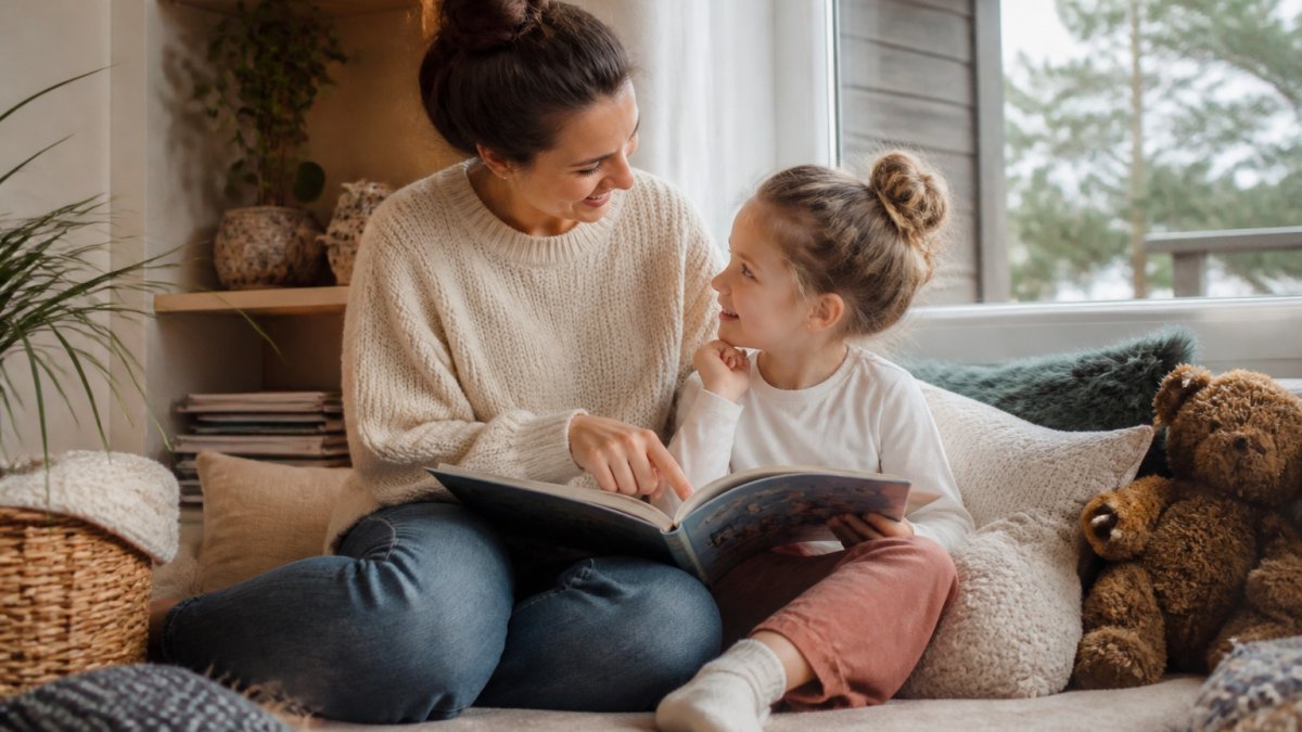 En voksen og et barn som sitter sammen i en koselig lesekrok hjemme, peker på en bildebok og ser på hverandre med varme og nysgjerrighet