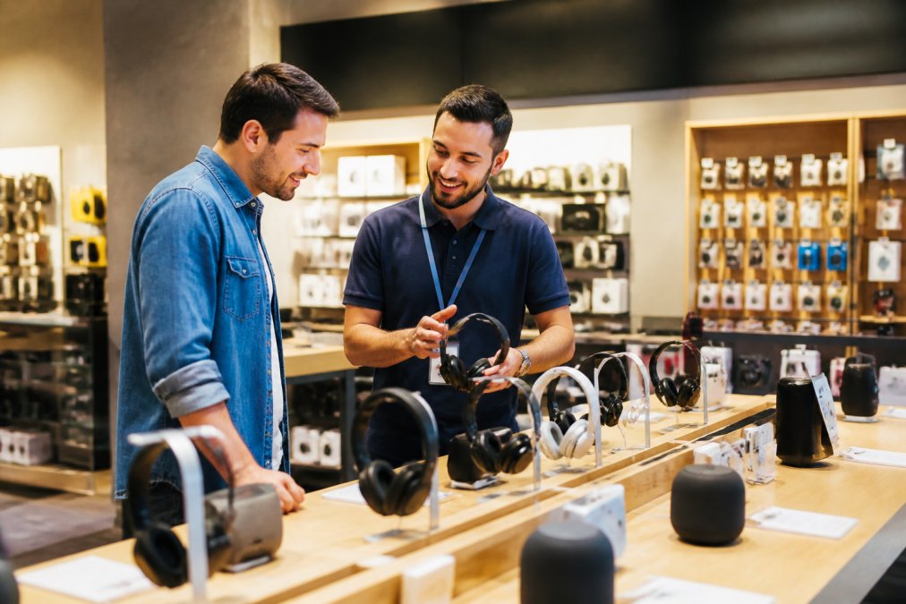 Kundeservice-medarbeider i en elektronikkbutikk hjelper en kunde med å velge riktig produkt, begge smilende og fokusert på en produktdisplay med moderne belysning