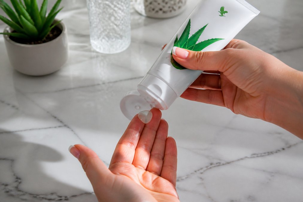 Hænder, der holder en Forever Aloe Vera Gelly-tube, med produktet, der bliver presset ud på fingerspidserne, hvid marmorbordplade i baggrunden, naturligt lys