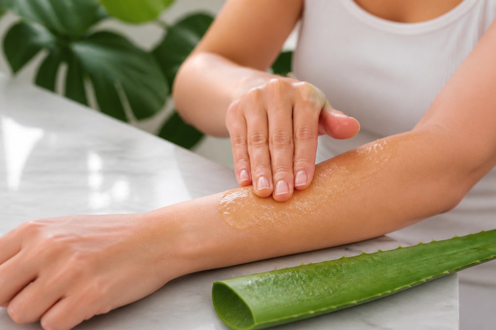 Close-up of a person gently applying clear aloe vera gel to their forearm, with a fresh aloe vera leaf placed beside them on a white surface