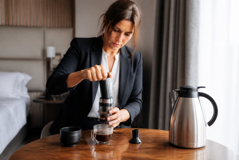 Travl forretningskvinde brygger manuel espresso på hotelværelset med fokuseret håndværk, manuel rejse espressomaskine og dampende kaffekop på træbordet med varmt vand i termokande