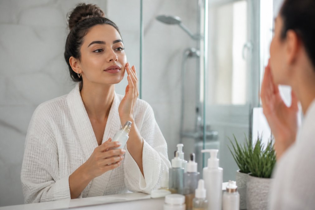 Person applying hydrating serums and moisturisers to face after treatment, with skincare products visible on bathroom counter in soft natural lighting