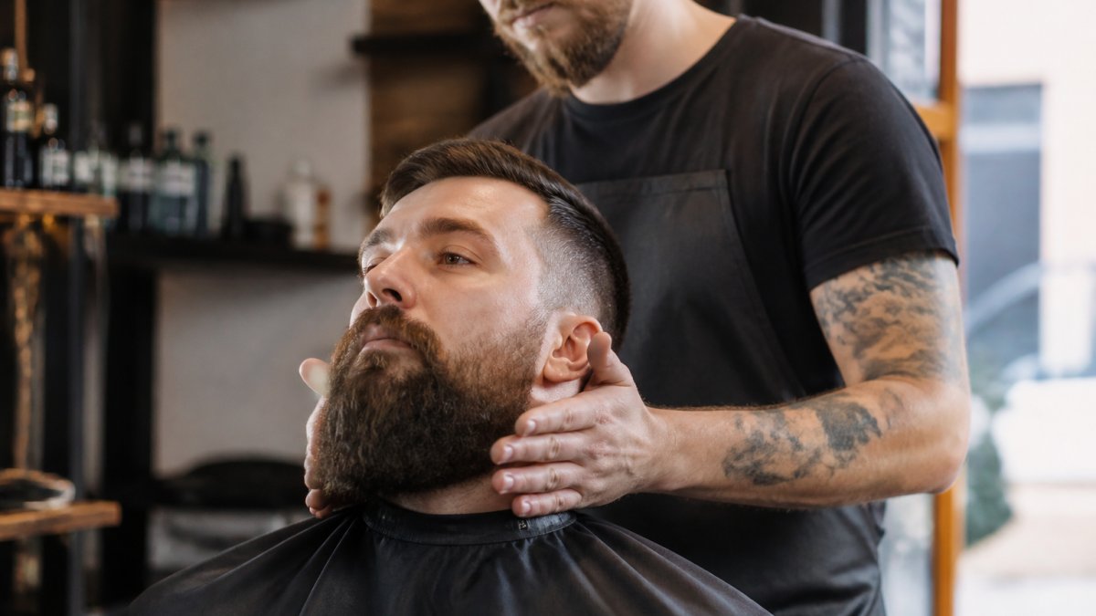 Professional barbier's hands demonstrating beard oil application technique, applying drops to fingertips and working oil through beard with downward strokes, natural morning light in modern barbershop