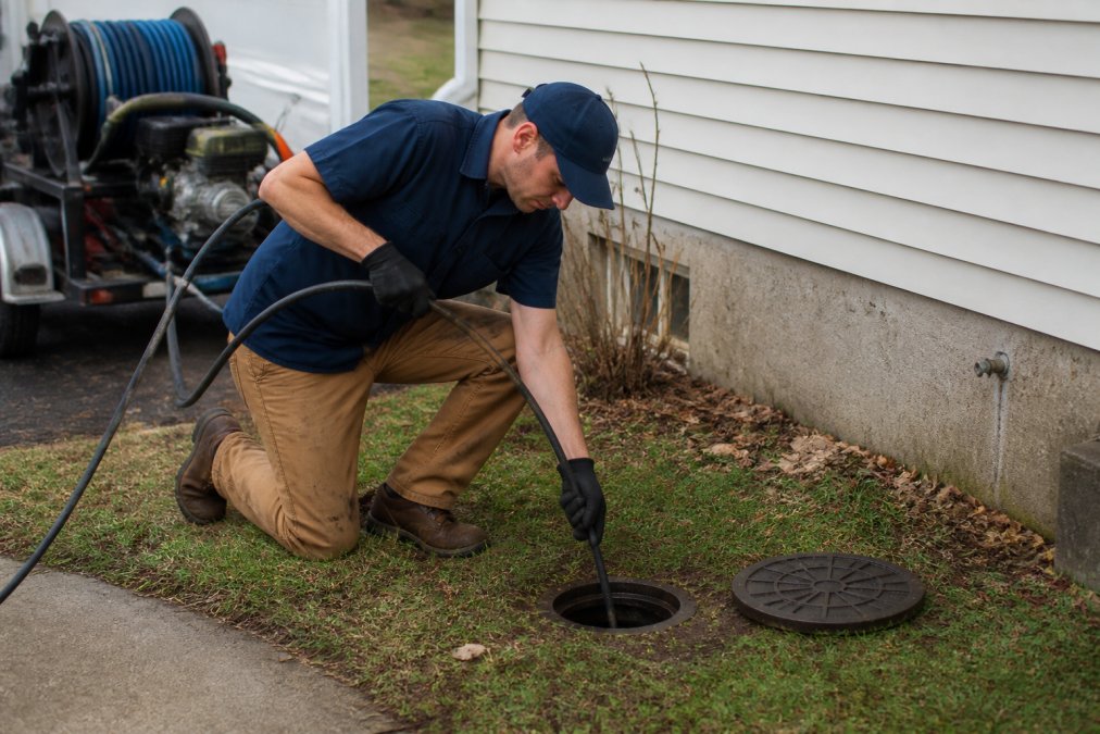 Professional plumber operating hydro-jetting equipment on a residential sewer line, directing the high-pressure water hose into the drain opening with focused intensity
