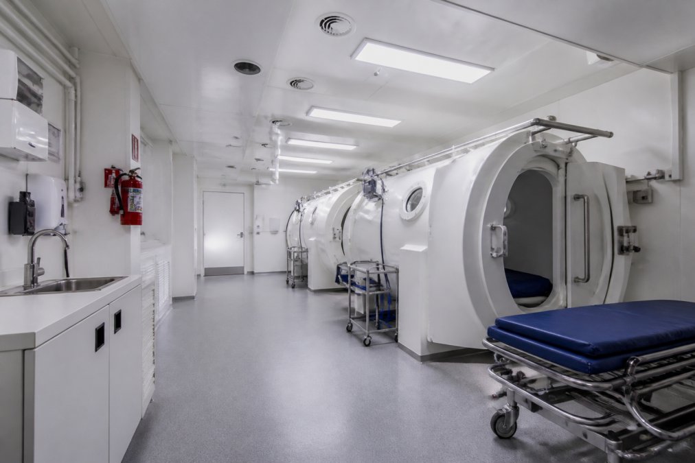 Modern hyperbaric chamber facility interior with clinical treatment environment, proper ventilation systems, safety signage, emergency equipment stations, and clean white walls with professional medical lighting