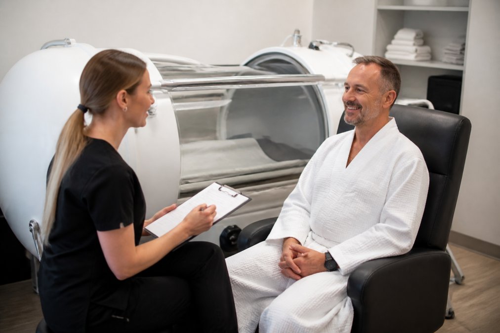 Healthcare technician conducting pre-session patient consultation and assessment next to a hyperbaric chamber in a clinical treatment room, with patient sitting and technician taking notes, natural medical facility lighting