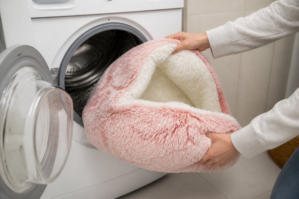 Hands placing a washable cat bed cover into a washing machine, showing the removable cover being loaded, bright laundry room lighting