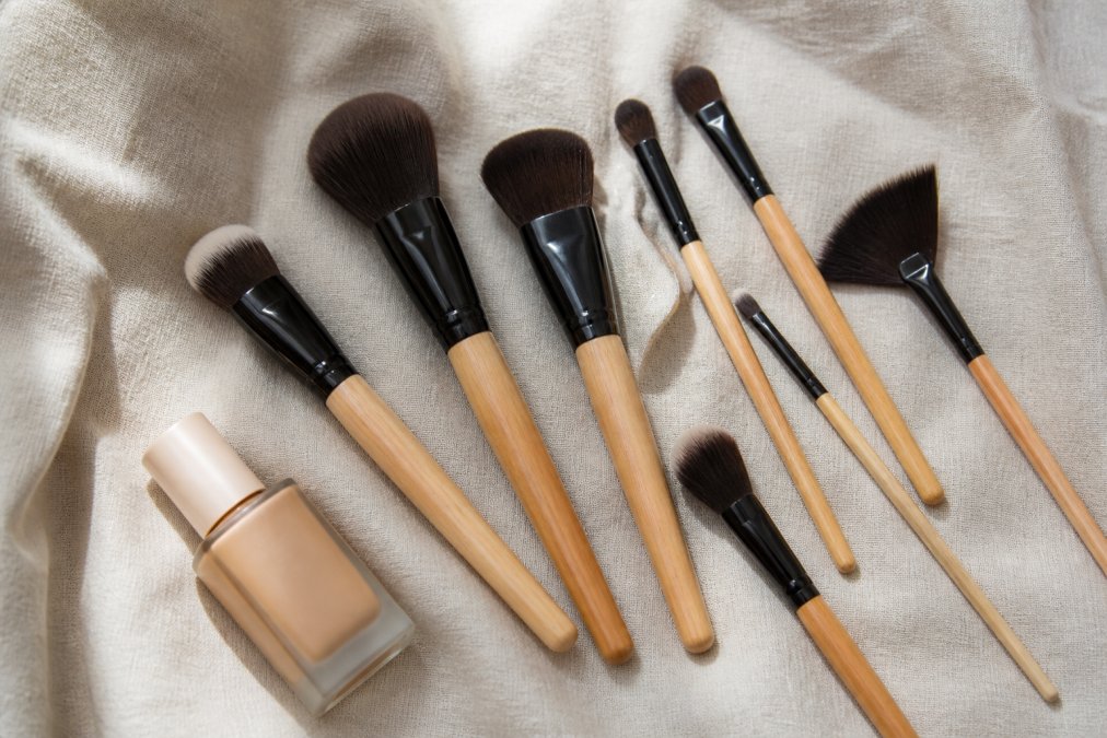 Flat lay of several professional makeup brushes with natural wood handles arranged neatly on a soft neutral linen background, with a small foundation product nearby