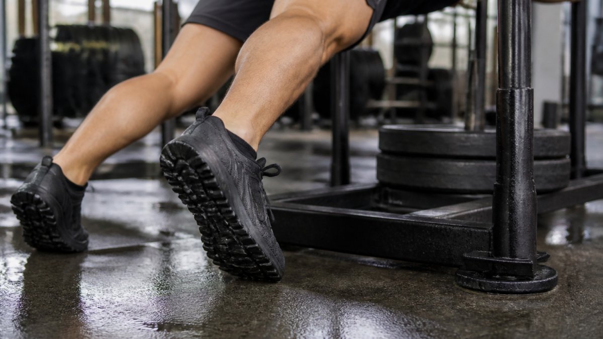 Close-up of an athlete's shoe sole gripping a metal sled platform during a push movement, showing deep tread patterns and foot positioning with sweat-dampened floor