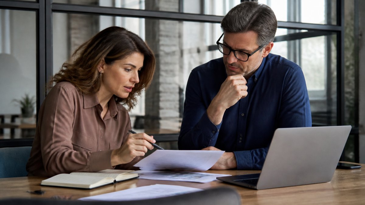Two business professionals reviewing documents and a laptop at a conference table, comparing vendor options in a bright office