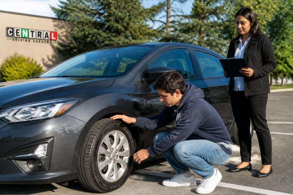 Student driver checking tire tread depth and sidewall condition on compact sedan in daylight, with vehicle clearly visible and examiner observing