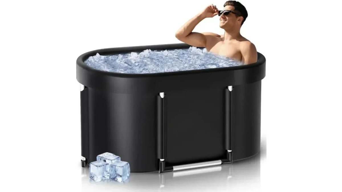 A compact foldable ice bath tub set up on a bathroom tile floor beside a folded towel and a small stool, natural window light, clean minimal apartment bathroom