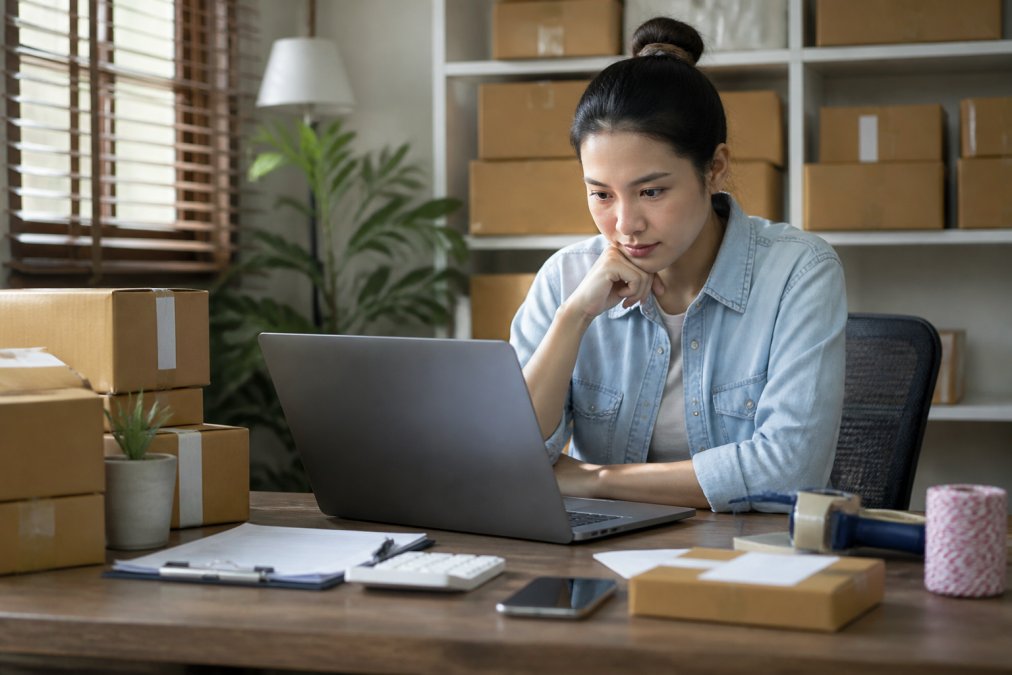 Small business owner or e-commerce manager at desk comparing shipping options on laptop, with shipping labels and packaging materials visible, natural office lighting