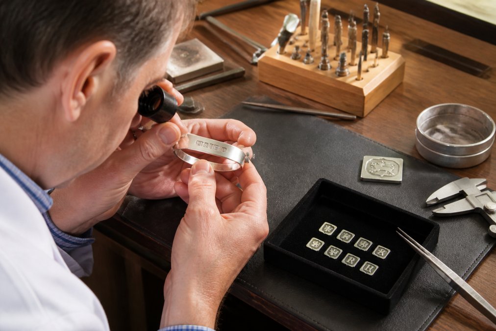 Jeweller using a magnifying glass to examine hallmarks on a silver piece on a workbench with professional lighting, carefully inspecting the sponsor's mark and assay office symbol