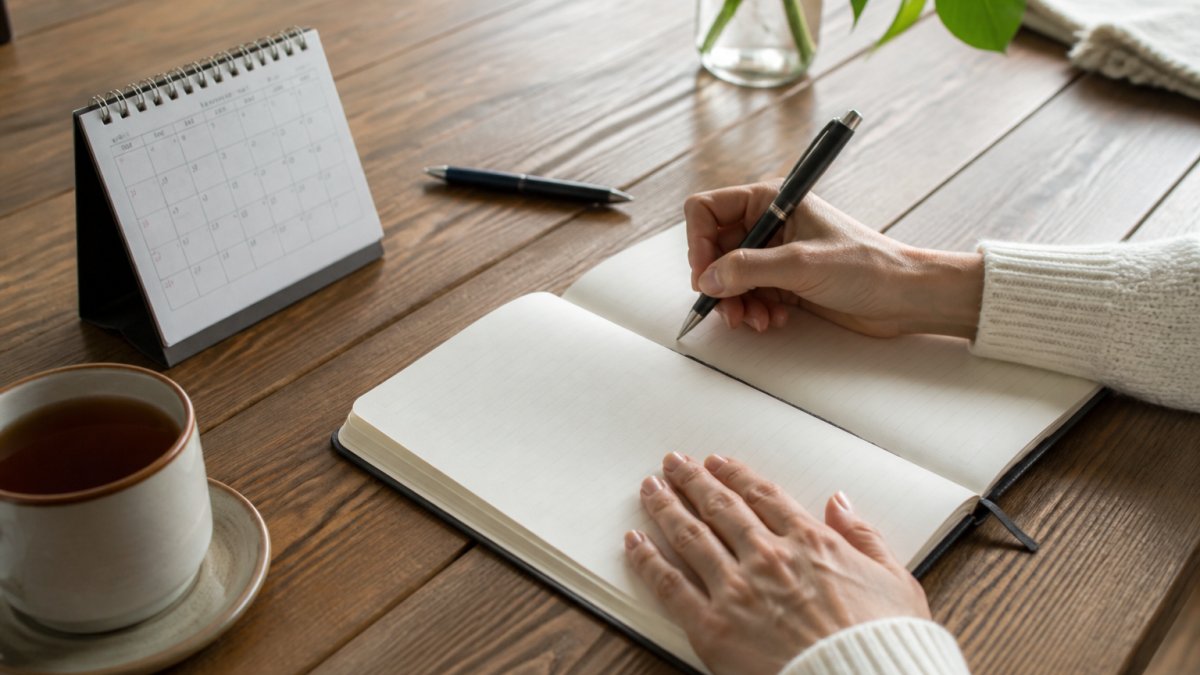 Person's hands writing in a journal at a wooden table with a cup of tea, calendar, and pen nearby, showing someone actively tracking daily health notes