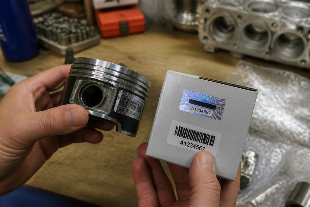 Close-up of hands holding an engine component beside its packaging, examining a holographic security label and barcode under bright workshop lighting, with the part number clearly visible on both the component and label