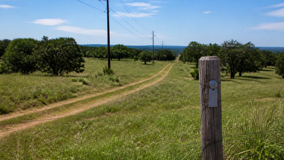 Rural property landscape showing visible utility poles and lines crossing pastureland, worn vehicle tracks indicating easement routes, and metal utility markers on fence posts in natural daylight