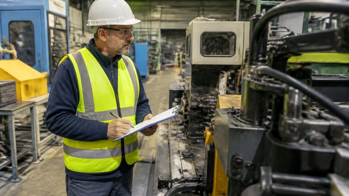 Safety manager conducting floor inspection in manufacturing facility, examining equipment and work areas with clipboard and checklist in hand