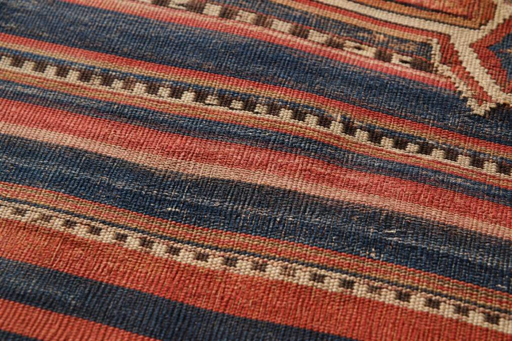 Close-up of antique kilim weave showing horizontal color striping and tonal variations in indigo and madder reds, natural sunlight highlighting the texture and subtle abrash pattern across warp threads