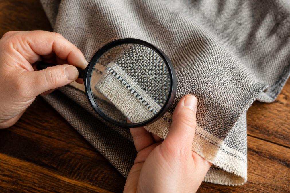 Magnifying glass inspecting woven fabric weaves to identify quality materials and selvage integrity.