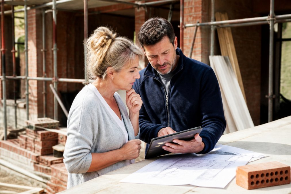 Huiseigenaar in gesprek met aannemer op een bouwplaats, beiden kijkend naar bouwplannen op een tablet, professionele en vriendelijke sfeer