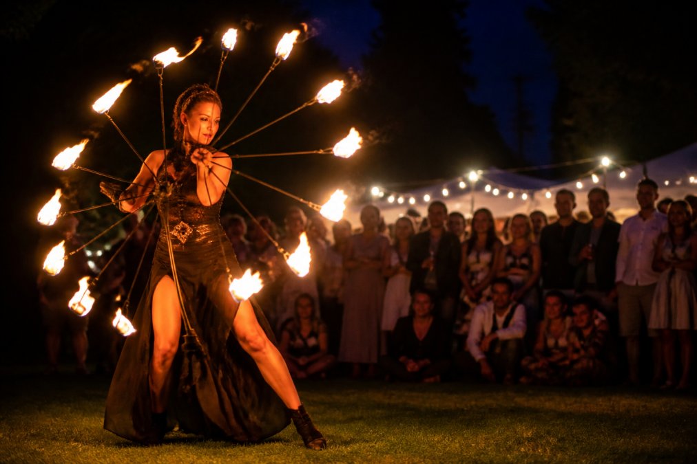 En ildartist spinner et flammende ildhjul i mørket foran et begejstret publikum ved en udendørs privat fest om aftenen, varmt orange lys fra flammerne reflekterer i publikums ansigter