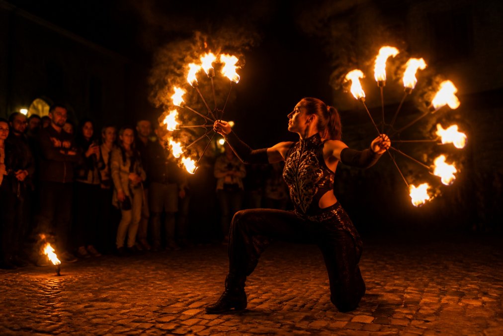 En performer med ildshow på en mørk scene, med flammende ildhjul og publikum i baggrunden, der ser fascineret på