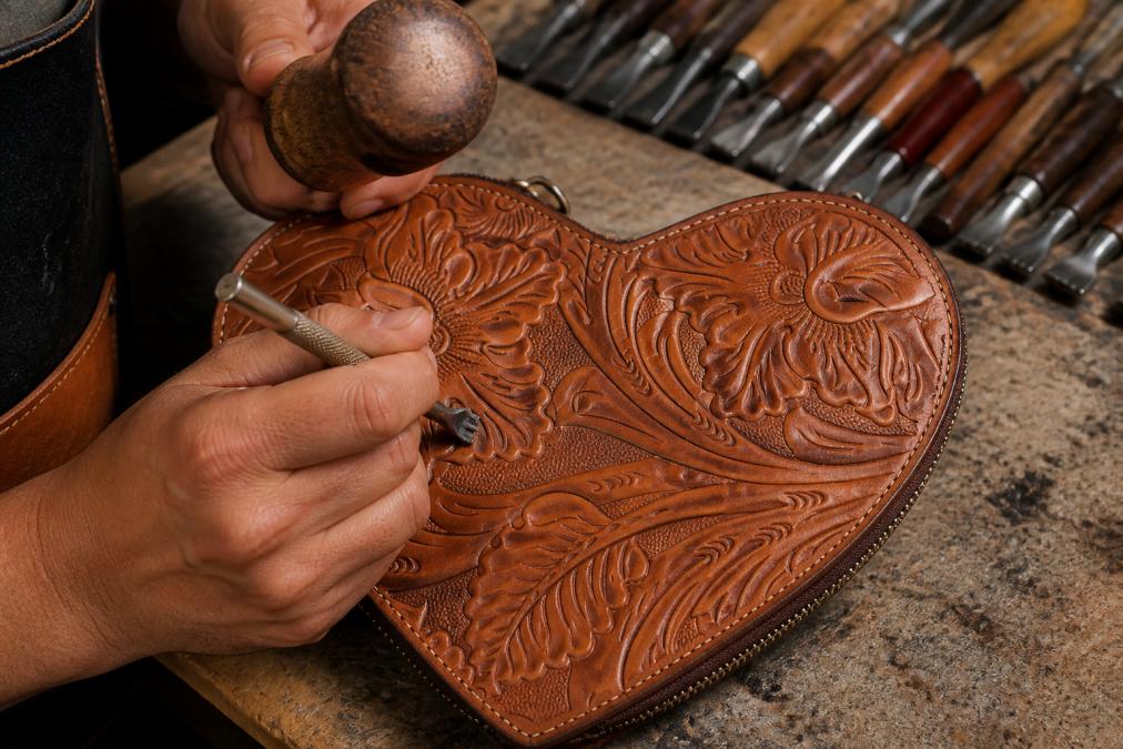 Close-up of artisan hands carefully carving intricate floral patterns into cognac-colored leather using traditional hand-tooling chisels and mallets, showing the precision and depth of the carved design in natural workshop lighting