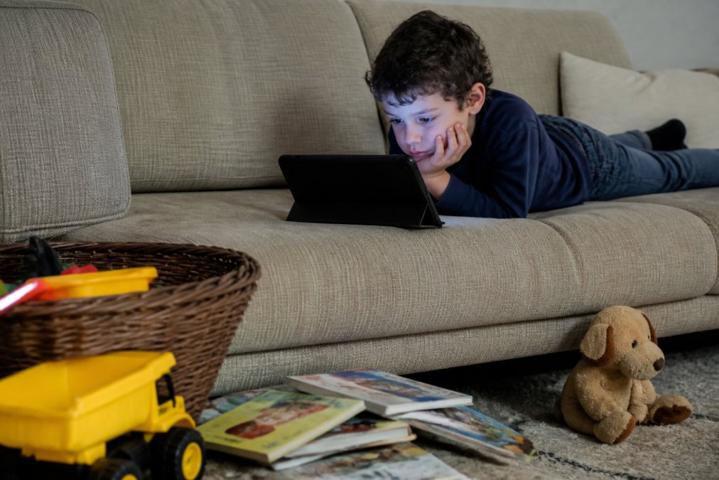 A young child lying on a couch staring blankly at a tablet screen, the glow reflecting on their face, with toys and books nearby untouched