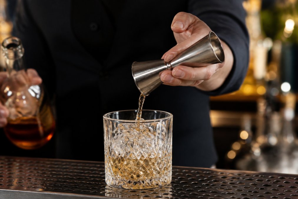 Close-up of a bartender's hands using a stainless steel jigger to measure spirits over a crystal rocks glass filled with ice, with professional bar lighting illuminating the precise measurement technique