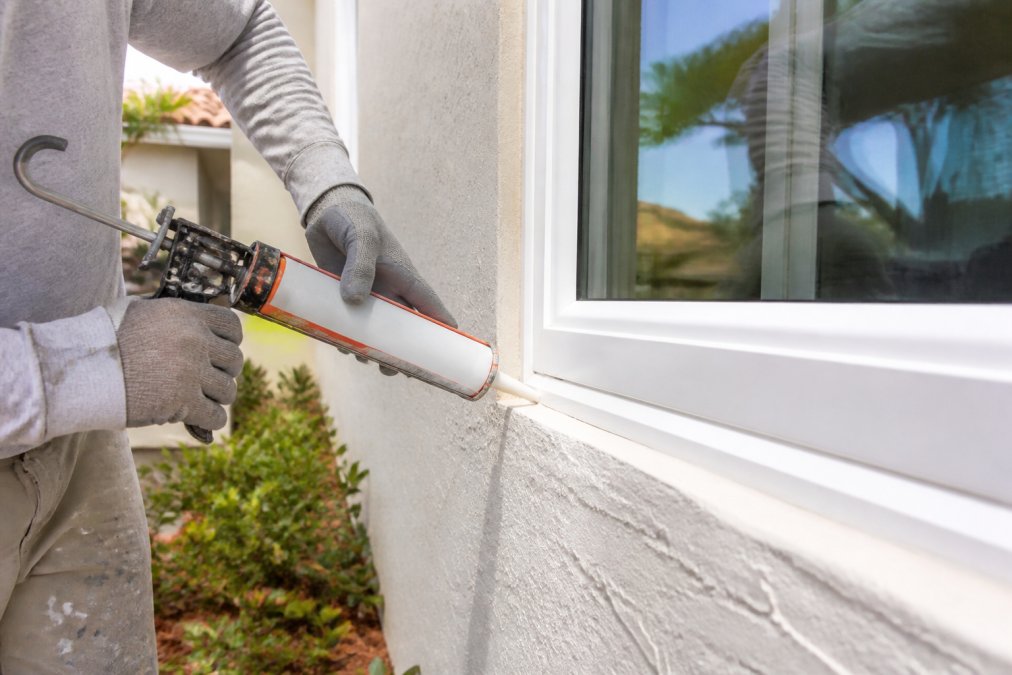 Close-up of a contractor's gloved hands applying weatherproof sealant in a continuous bead around the frame of a newly installed impact window on a sunny residential exterior, with caulking gun and clean white frame visible
