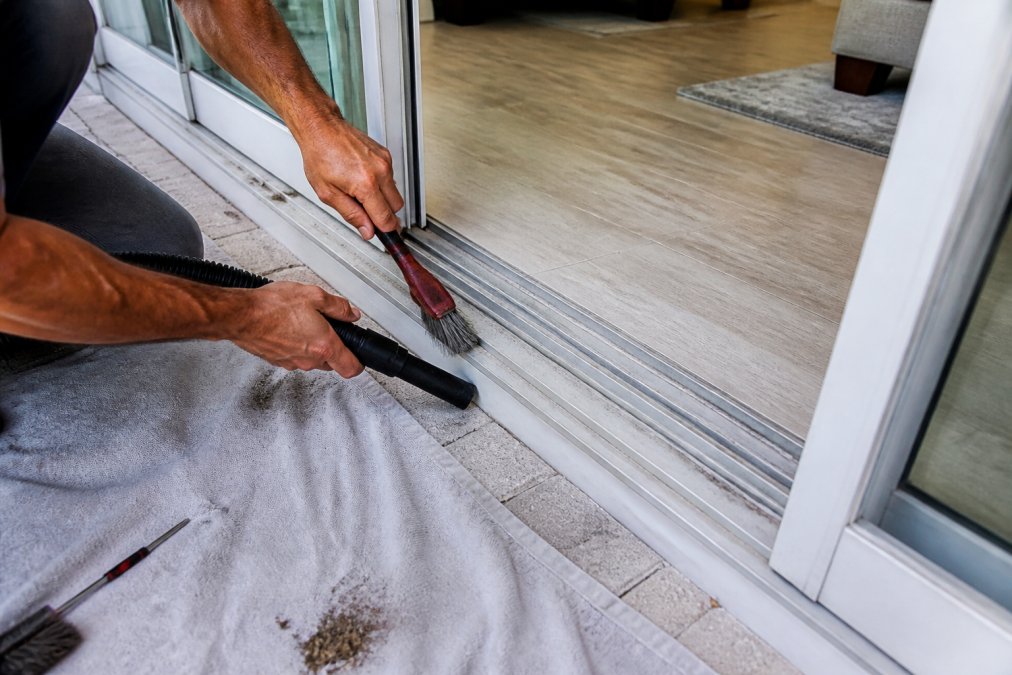 A homeowner using a stiff brush and vacuum to clean debris from a sliding door track during maintenance.