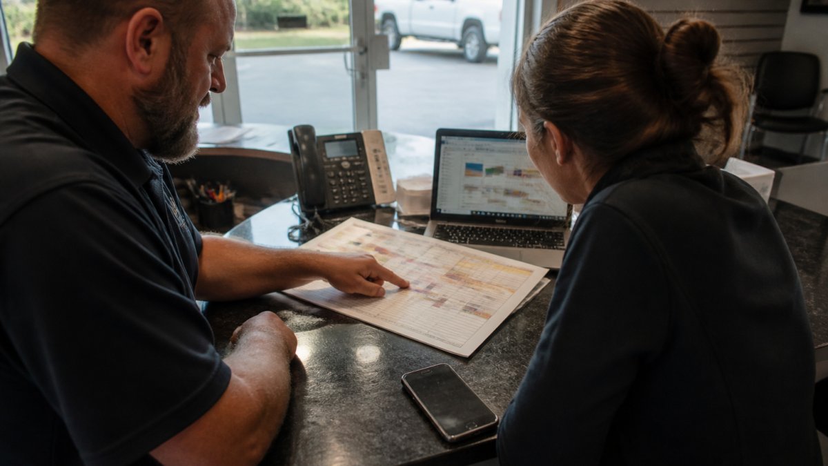 A home service business owner and a team member reviewing a printed workflow map and laptop screen at a front desk, with a phone and scheduling calendar visible