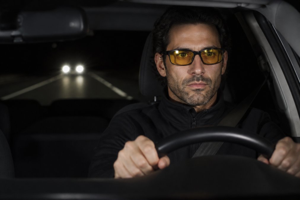 Close-up of a driver at night looking ahead at the road with oncoming headlights visible in the distance, wearing specialized night-driving glasses with subtle yellow tint, hands on steering wheel